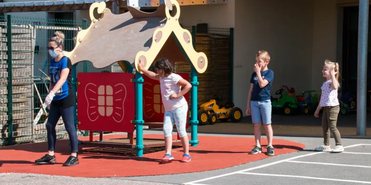 Une animatrice, masque et gants, surveille des enfants sur un terrain de jeu. Le directeur ACM veille à la sécurité.