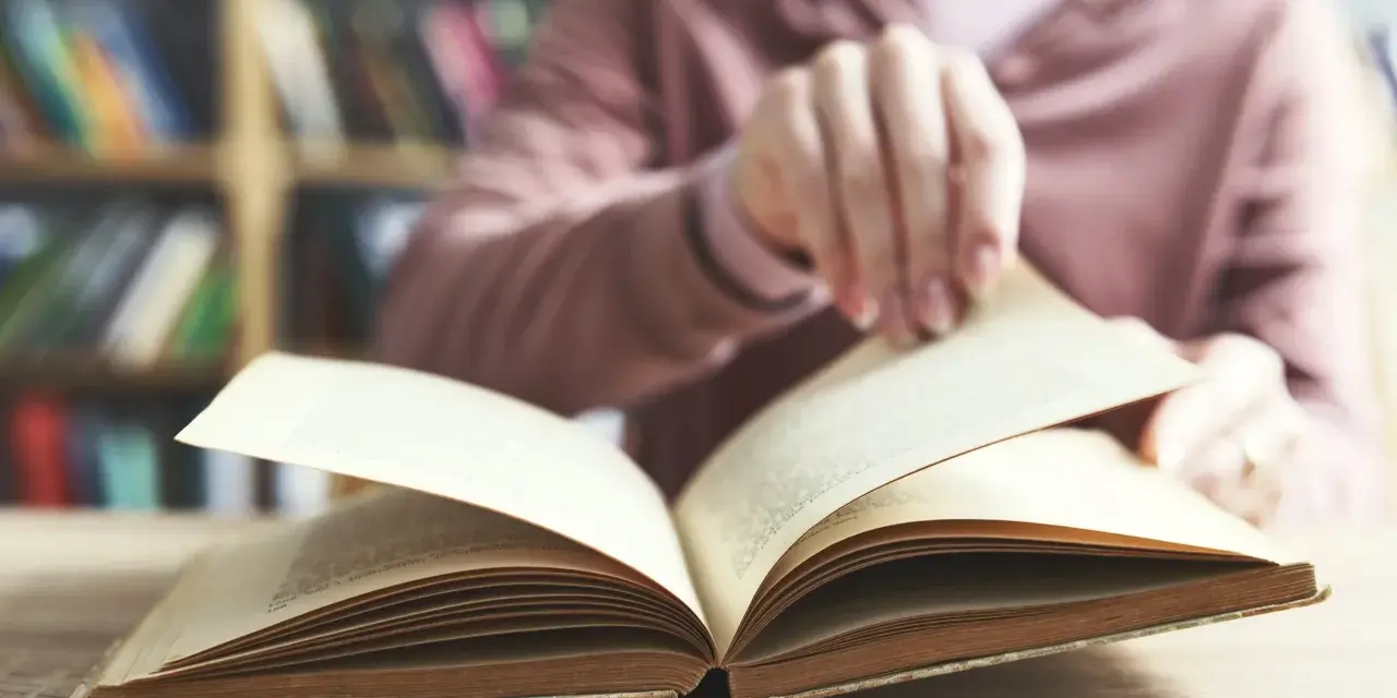Otwarta księga na stole, w tle regały pełne książek. To początek podróży przez arcydzieła literatury światowej, lista skarbów do odkrycia.