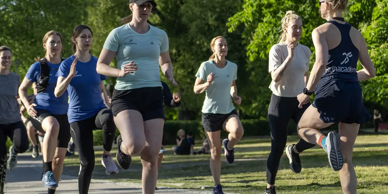 En grupp kvinnor tränar löpning utomhus. De springer i ett parkliknande område, redo att springa 1 km tid.