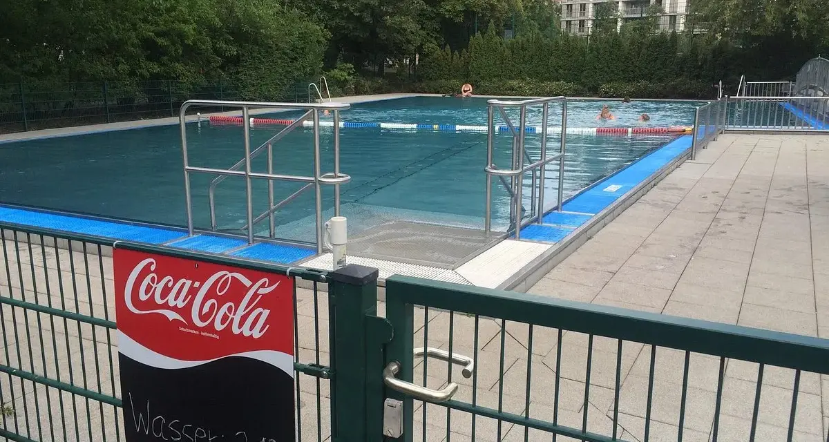 Erfrischung im Monbijou Bad: Wasser 21°C. Menschen schwimmen im blauen Becken, umgeben von Bäumen und einem modernen Gebäude.