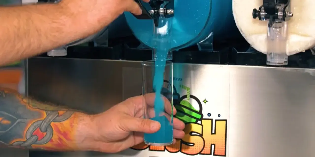 Ein Arm mit einem Ketten-Tattoo füllt ein Glas mit blauem Slush Eis aus einem Automaten.