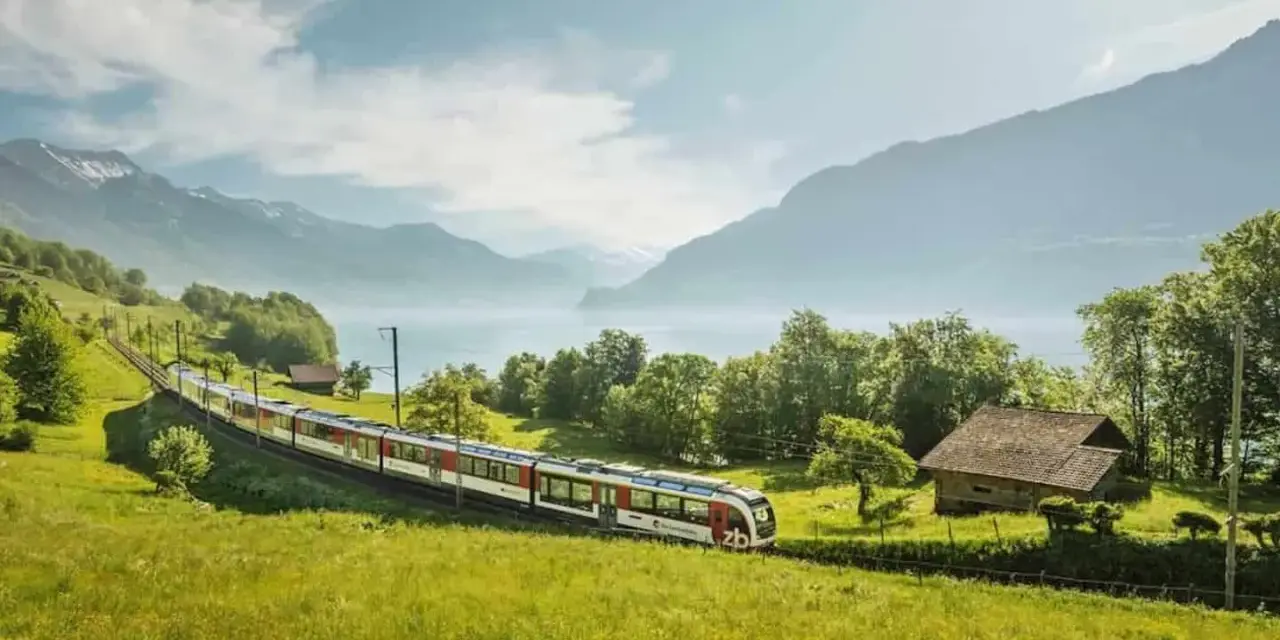 A train travels through a lush green valley, with a lake and mountains in the background. This scenic route from Lucerne to Montreux offers breathtaking views.