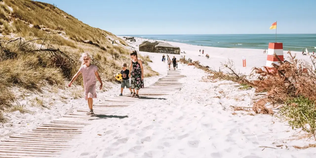 Familj promenerar på en strand med vit sand i Danmark. En livräddartorn och bunkrar syns i bakgrunden.