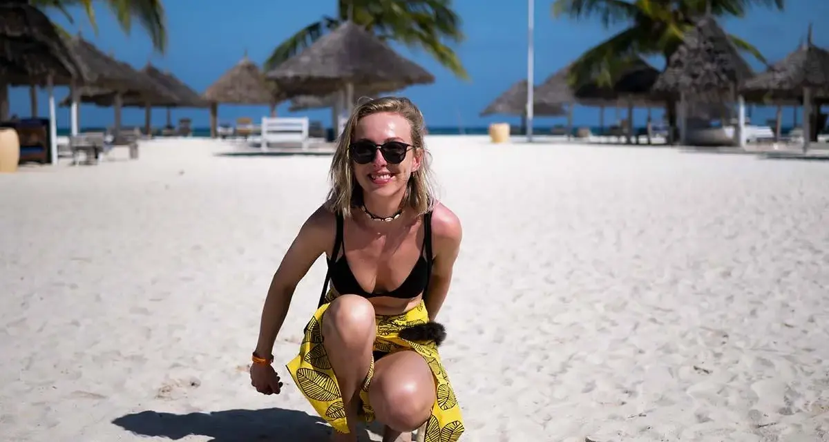 Uśmiechnięta blondynka w bikini i żółtej spódniczce kuca na piaszczystej plaży na Zanzibarze. W tle palmy i słomiane parasole.