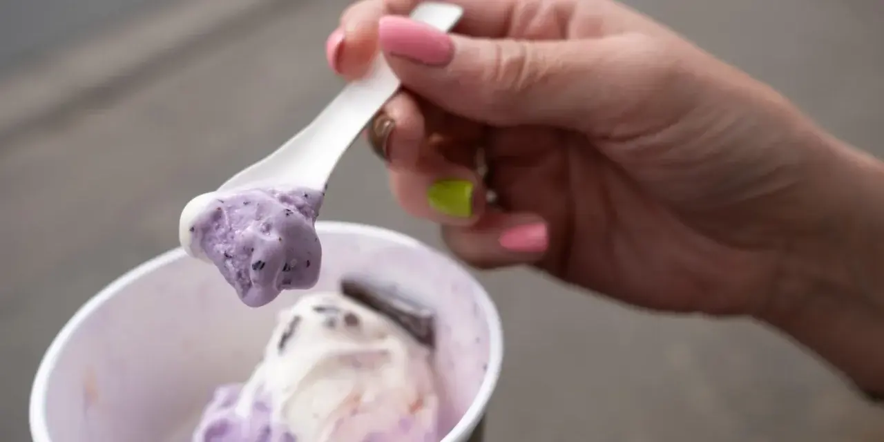 Eine Hand mit pink lackierten Nägeln hält einen Löffel mit zwei Kugeln Eis im Becher, italienisch.