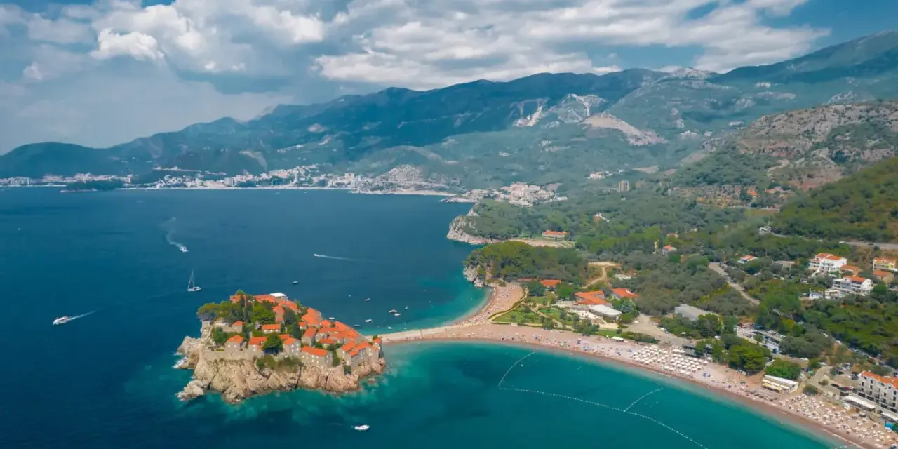 Sveti Stefan, Czarnogóra czy warto? Widok z lotu ptaka na malowniczą wyspę połączoną z lądem, turkusowe morze i górzyste wybrzeże.