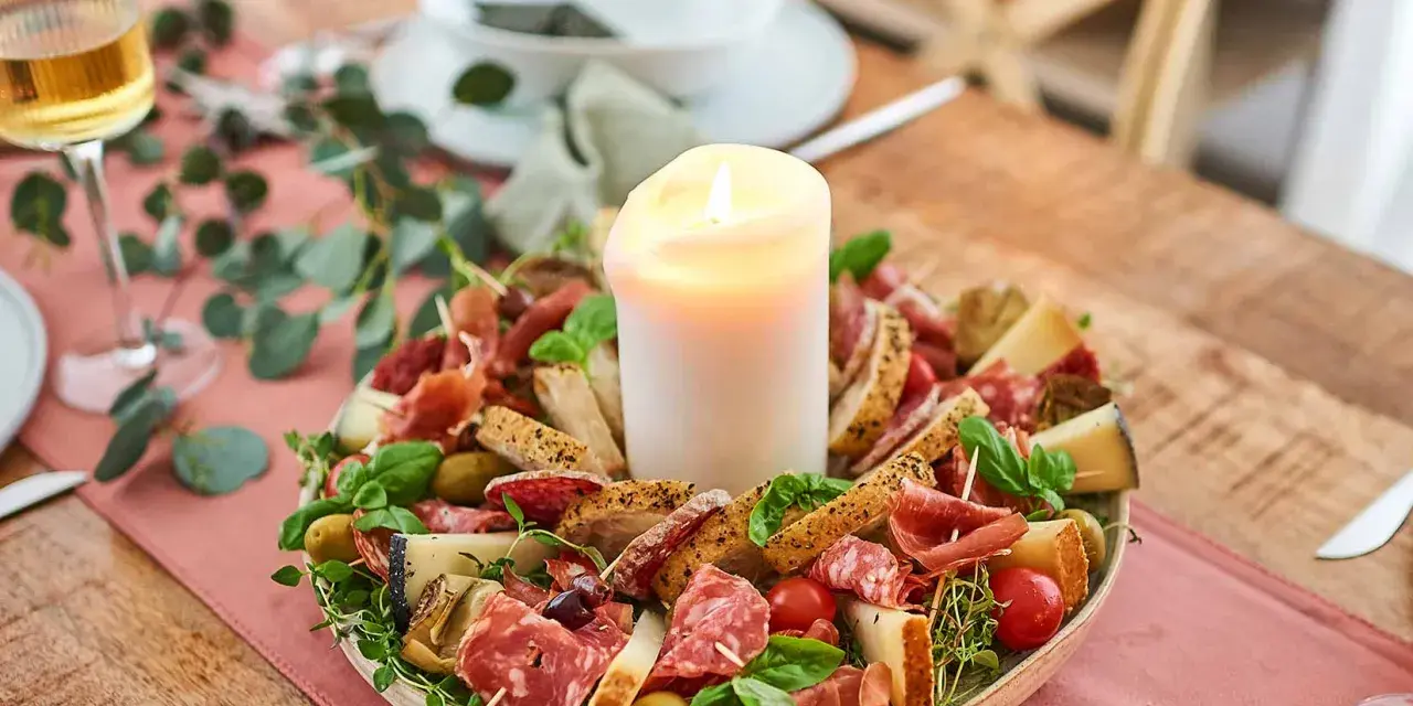 Antipasti selber machen: Eine festliche Platte mit Wurst, Käse, Brot, Oliven und Tomaten, umgeben von grünen Zweigen, mit einer Kerze in der Mitte.