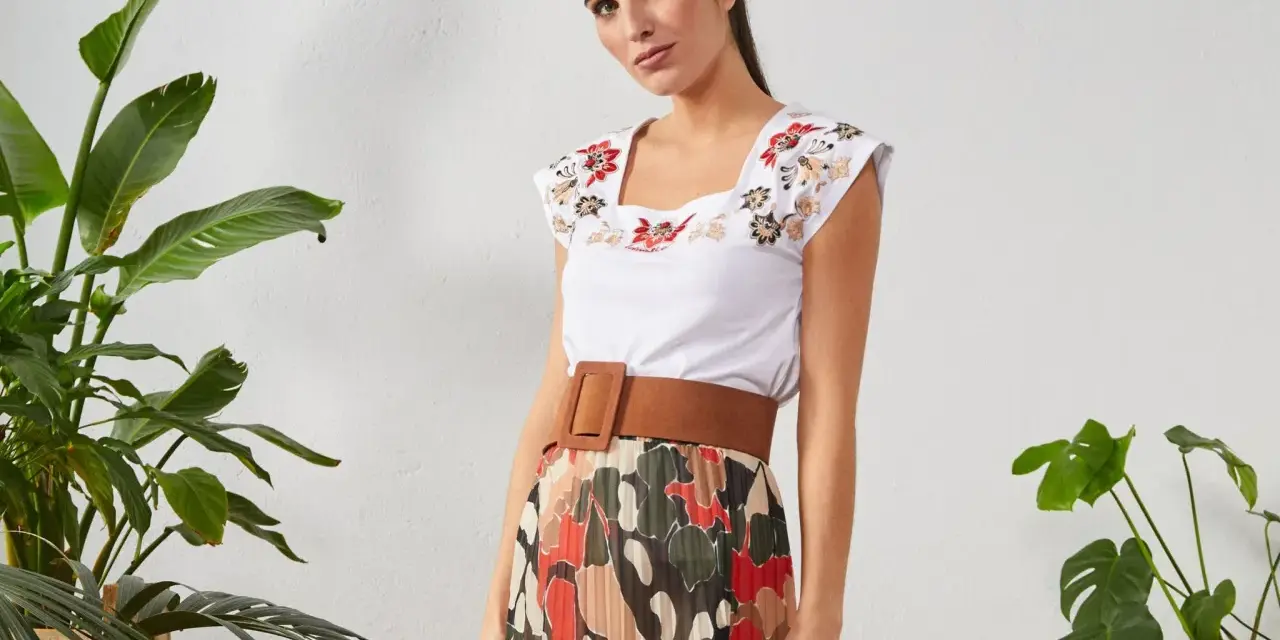 Mujer con top blanco bordado y falda plisada de estampado abstracto. Un cinturón marrón ancho realza la cintura.