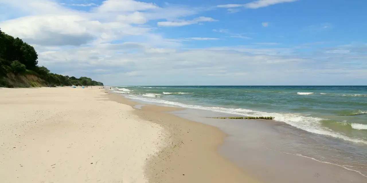 Plaża nad morzem, gdzie fale delikatnie obmywają piasek. W oddali widać zalesione wzgórze, a na niebie chmury. Idealne miejsce na spacer dla gąsek.