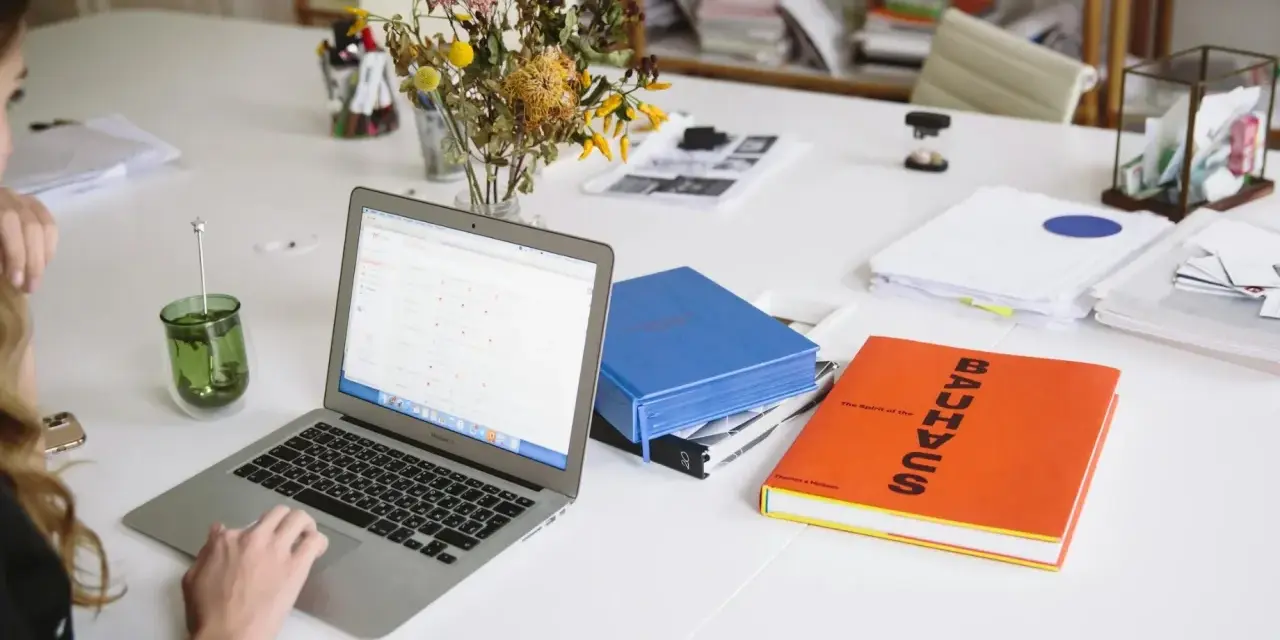Escritorio con portátil, libros y flores. Un espacio de trabajo que parece una copistería por la cantidad de documentos y la organización.