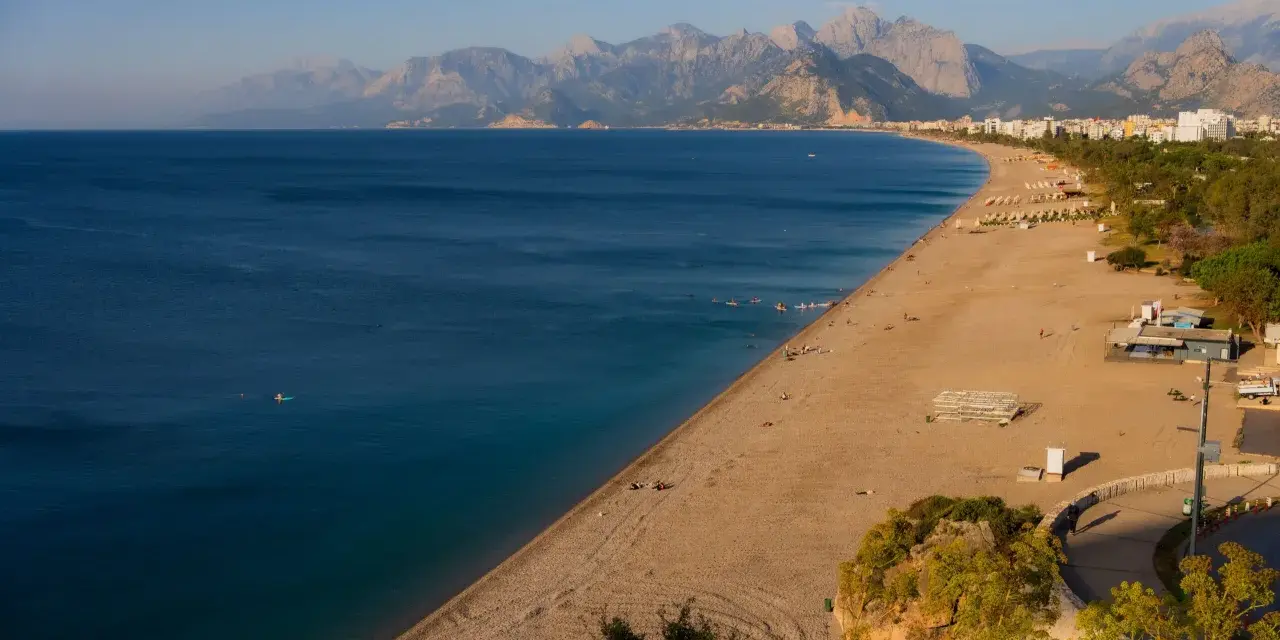 Słoneczna plaża na riwierze tureckiej z turkusowym morzem i górami w tle. Kilka osób pływa, inni odpoczywają na piasku.