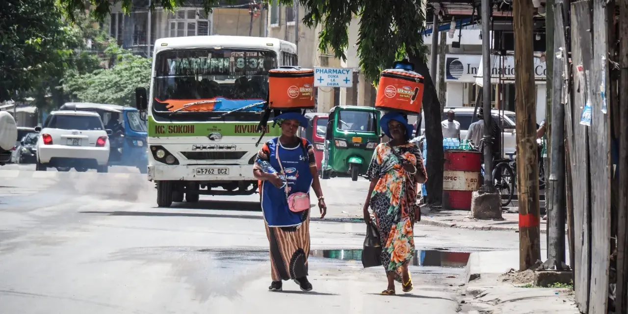 Dwie kobiety niosą na głowach pomarańczowe lodówki w upalny dzień w Dar es Salaam. W tle autobus i ruchliwa ulica.
