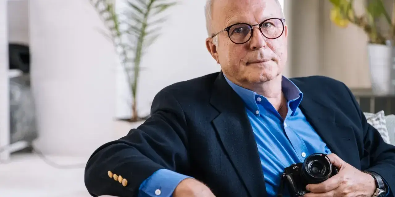 Tomaszewski fotograf w okularach, w granatowej marynarce i niebieskiej koszuli, trzyma aparat.