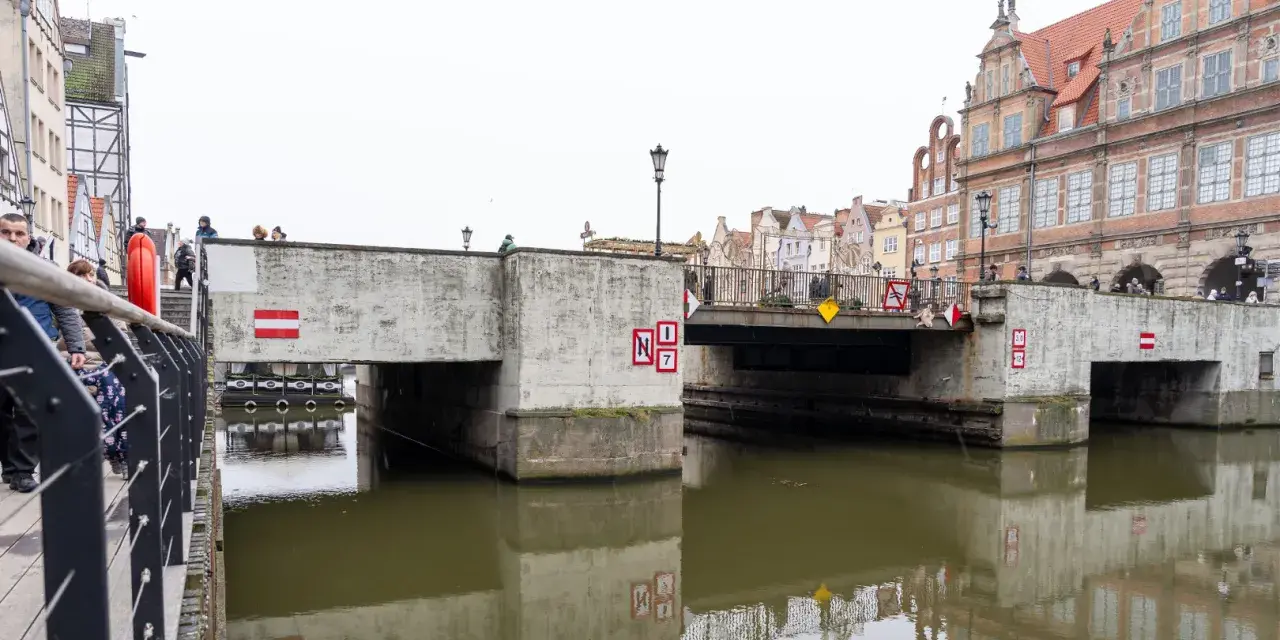 Nad kanałem w Gdańsku wznosi się betonowy zielony most, ozdobiony czerwono-białymi znakami. Ludzie spacerują po jego balustradzie.