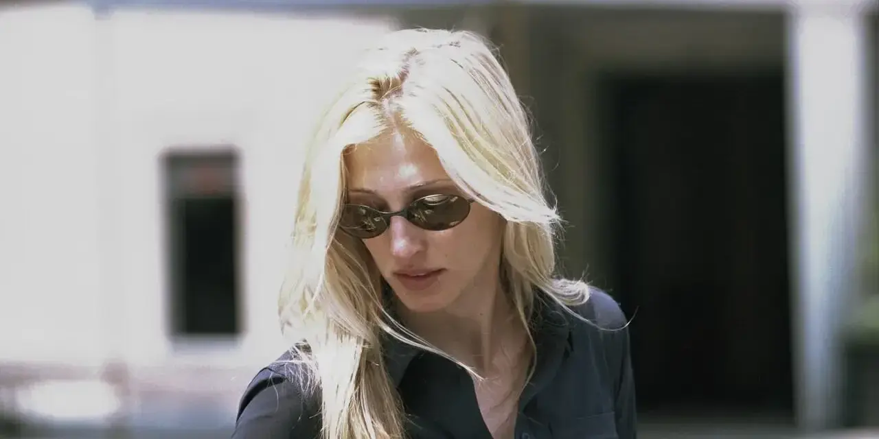 Blondynka w okularach przeciwsłonecznych, w czarnej koszuli, emanuje klasycznym, ponadczasowym stylem Carolyn Bessette.