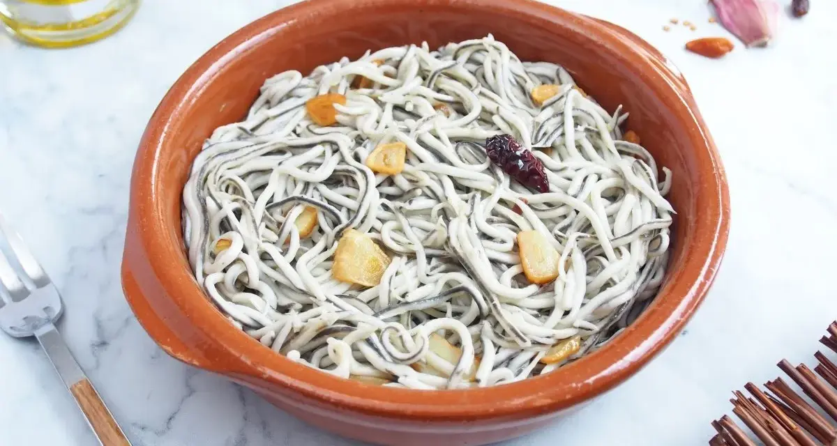 Un cuenco de barro con gulas al ajillo, adornado con láminas de ajo dorado y un chile seco.