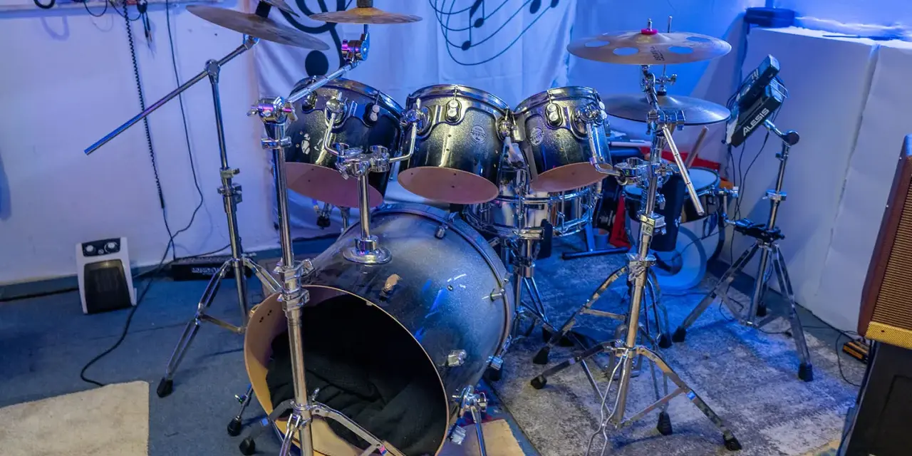 Der **Aufbau Schlagzeug** ist bereit für den nächsten Gig. Glänzende Trommeln und Becken stehen auf stabilen Stativen, beleuchtet von blauem Licht.