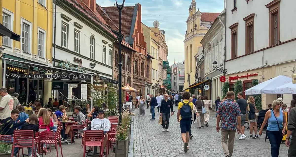 Tłum ludzi spaceruje brukowaną ulicą w Wilnie, siedząc przy stolikach restauracyjnych. Czy wyjazd na Litwę jest bezpieczny? Wygląda na to, że tak!