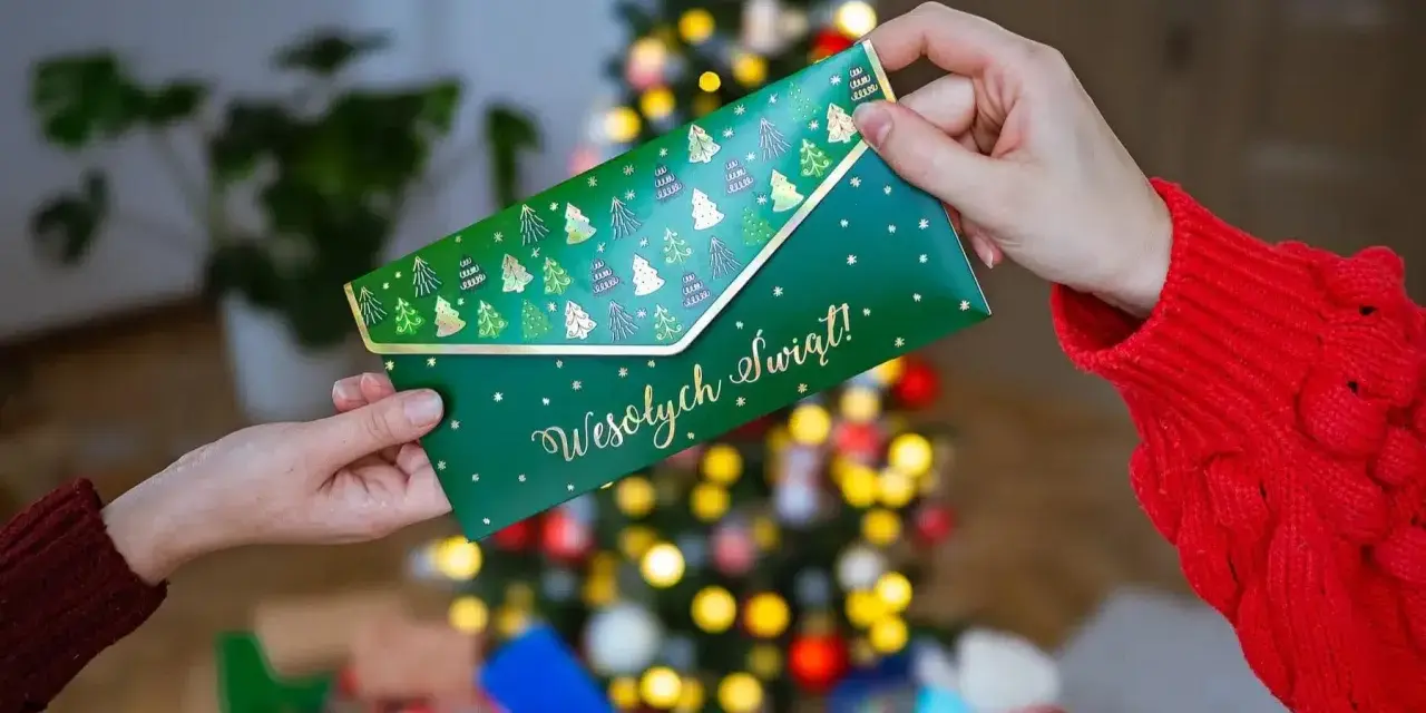 Jak zapakować voucher na prezent? Zielona koperta z choinkami i napisem "Wesołych Świąt!" jest przekazywana w świątecznej atmosferze.