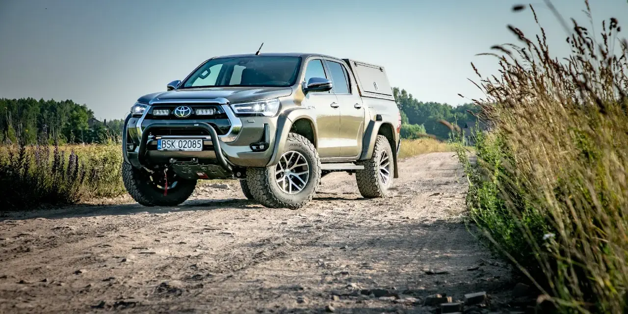 Złota toyota terenowa z wyciągarką i zabudową pędzi po polnej drodze, otoczona dziką roślinnością.
