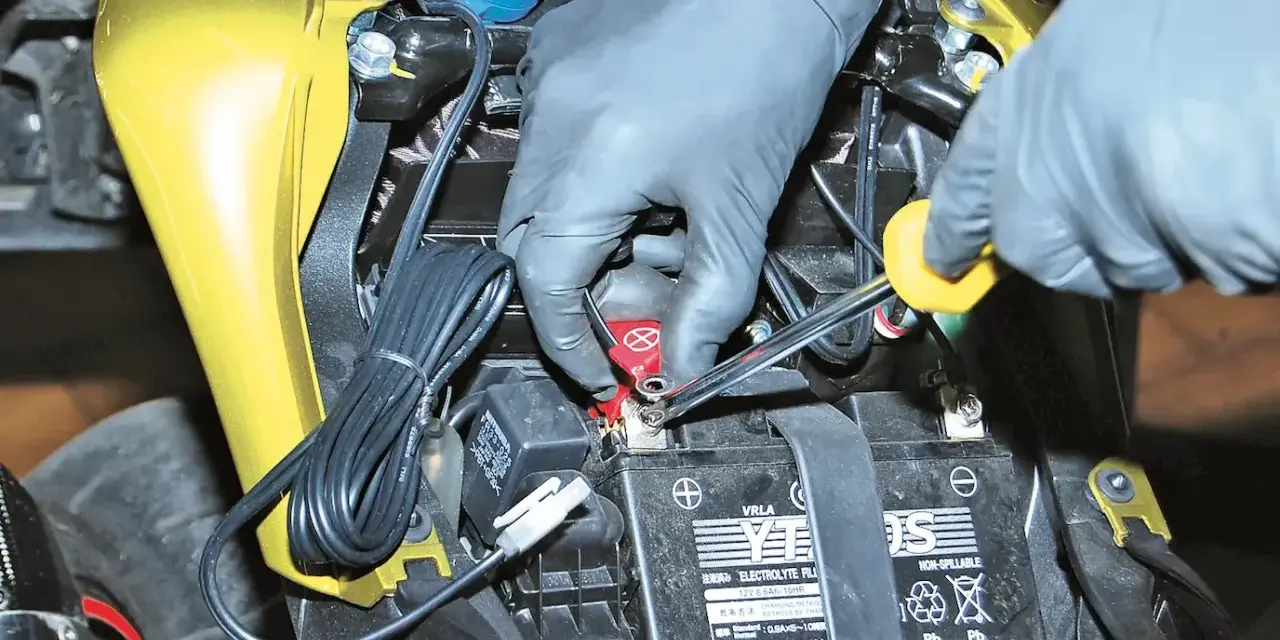 Mani guantate usano una chiave per staccare la batteria moto, scollegando il cavo rosso.