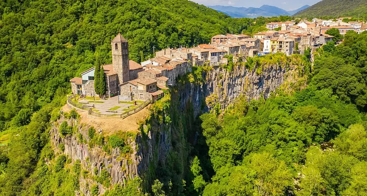 Un borgo medievale, Castellfollit de la Roca, aggrappato a una scogliera basaltica, circondato da lussureggianti foreste verdi.