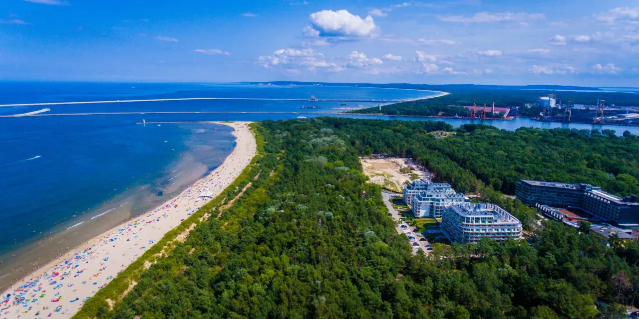 Widok z lotu ptaka na najszerszą plażę w Polsce, pełną wypoczywających ludzi, otoczoną lasem i nowoczesnymi budynkami.