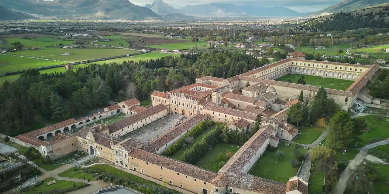 Vasta Certosa di Padula, un complesso monastico imponente con cortili verdi, circondato da alberi e montagne sullo sfondo.