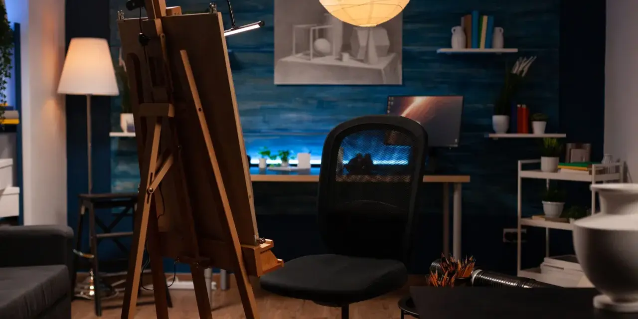 Un estudio de pintura en casa con caballete, silla de oficina, lámpara de pie y un escritorio con ordenador.