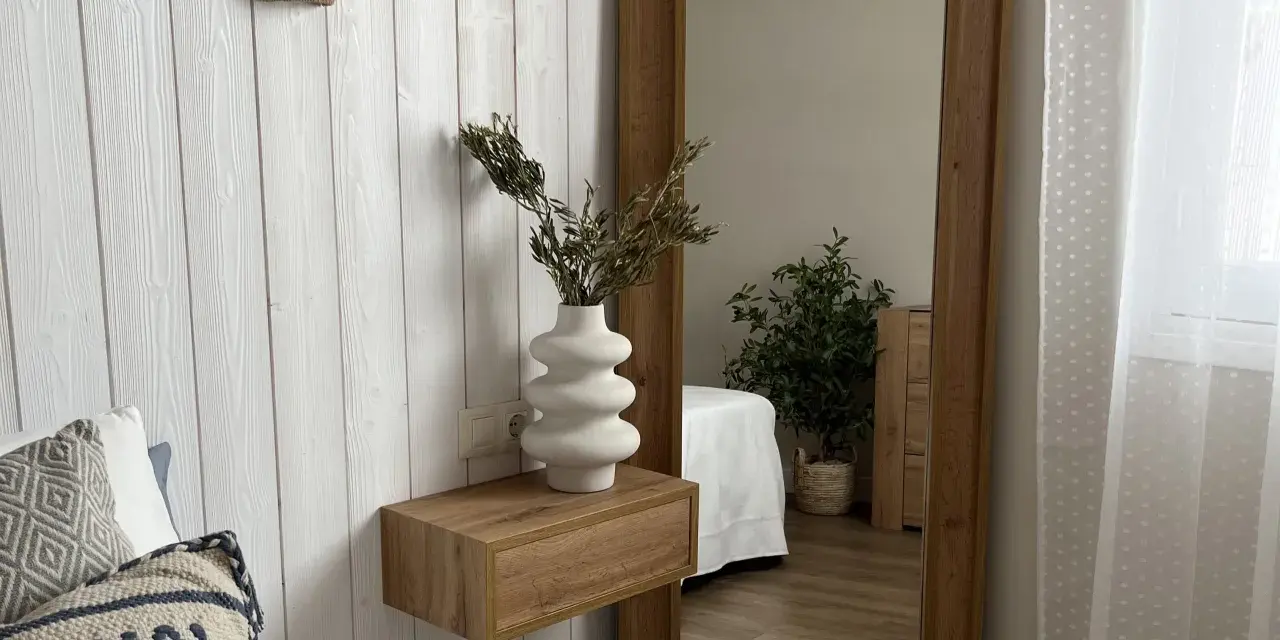 Estantería flotante de madera con jarrón blanco y ramas secas, ideal para ideas para decorar rincones pequeños. Reflejo de planta y cama en espejo grande.