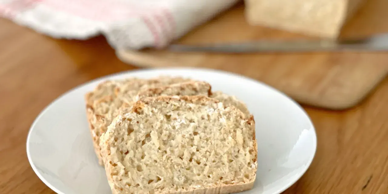 Skivor av hembakat bröd på en vit tallrik, perfekt för snabb bakning. En bit bröd ligger på en skärbräda i bakgrunden.