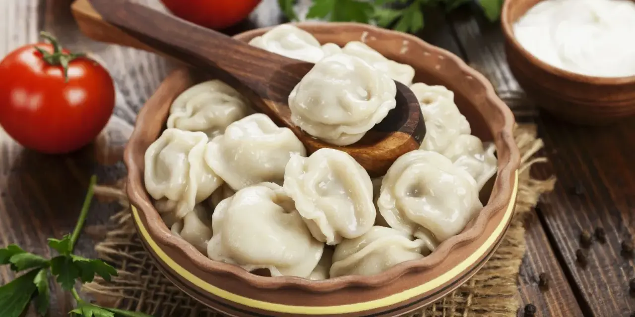 Pielmieni co to? Pyszne, gotowane pierożki w glinianej misce, z drewnianą łyżką, pomidorami i śmietaną w tle.