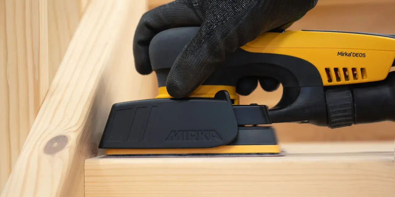 En hand med handske slipar en träyta med en Mirka DEOS-slipmaskin. Perfekt för att slipa borr och andra trädetaljer.