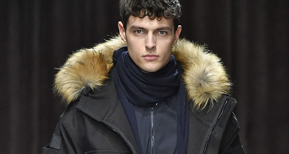 Modello con giacca Hugo marca con cappuccio bordato di pelliccia, sciarpa blu scuro, sguardo intenso.