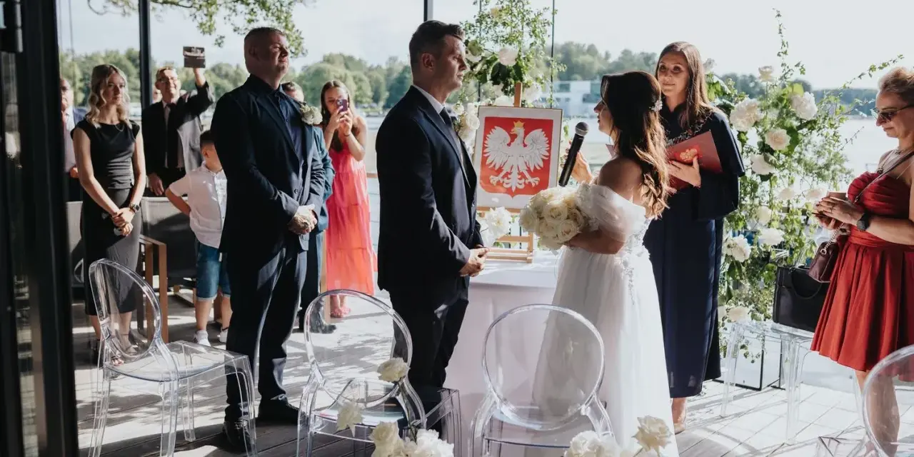 Uroczyste wesele w ogrodzie. Para młoda stoi przed urzędniczką, goście obserwują ceremonię.