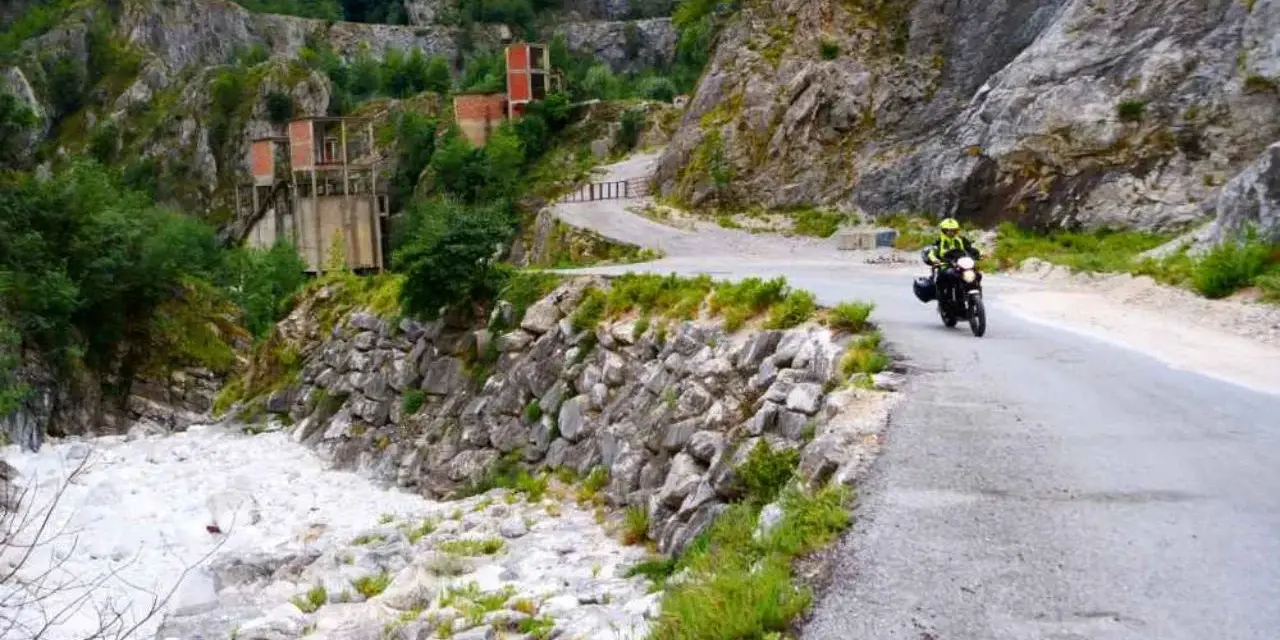 Un motociclista percorre una strada tortuosa tra le Alpi Apuane, con un fiume di sassi e ruderi industriali sullo sfondo.