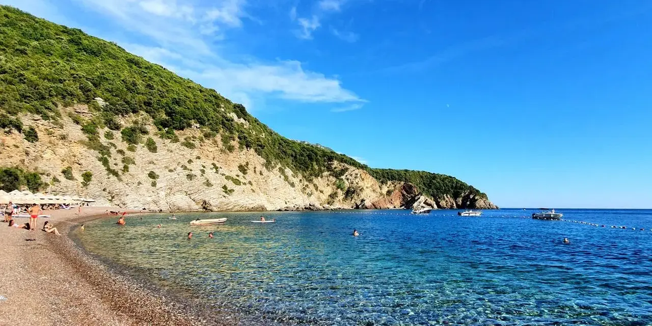 Słoneczne plaże w Budvie, z turkusową wodą i zielonymi wzgórzami w tle. Ludzie relaksują się na kamienistym brzegu i pływają w morzu.