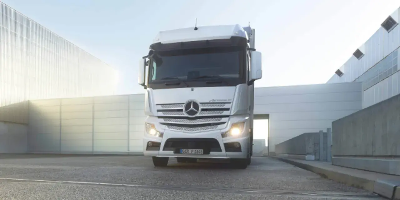 Biały Mercedes Actros MP4 z włączonymi światłami stoi przed nowoczesnym budynkiem.