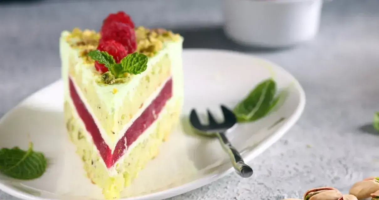 Pyszny tort pistacjowy z malinowym nadzieniem, udekorowany malinami i miętą. Obok leżą łuskane pistacje.