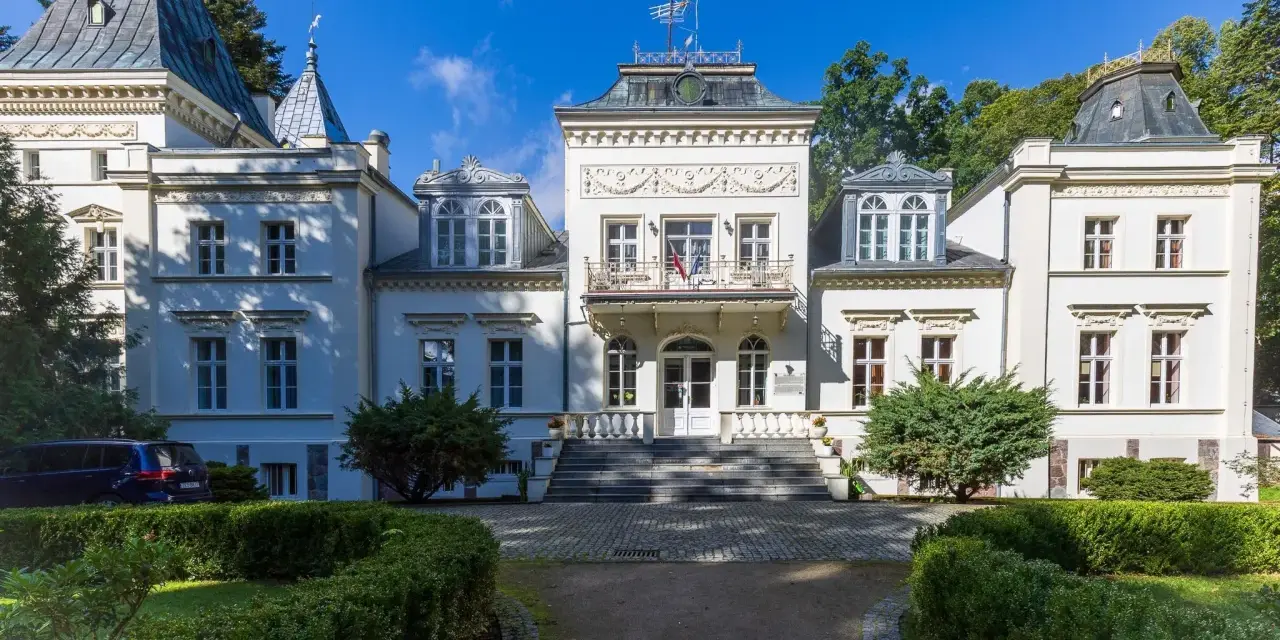 Elegancki pałac w Nosowie z wieżyczkami i ozdobnym balkonem, otoczony zielenią.