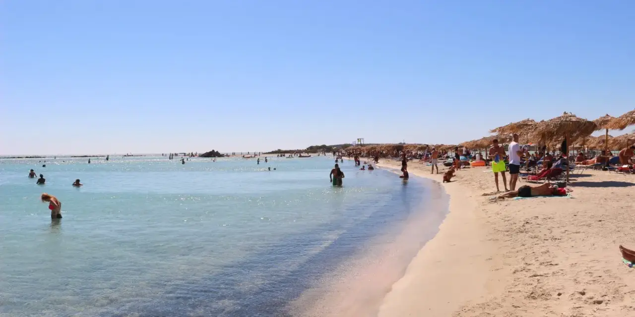 Słoneczna plaża Elafonisi z turkusową wodą i piaskiem, pełna ludzi kąpiących się i odpoczywających pod parasolami.