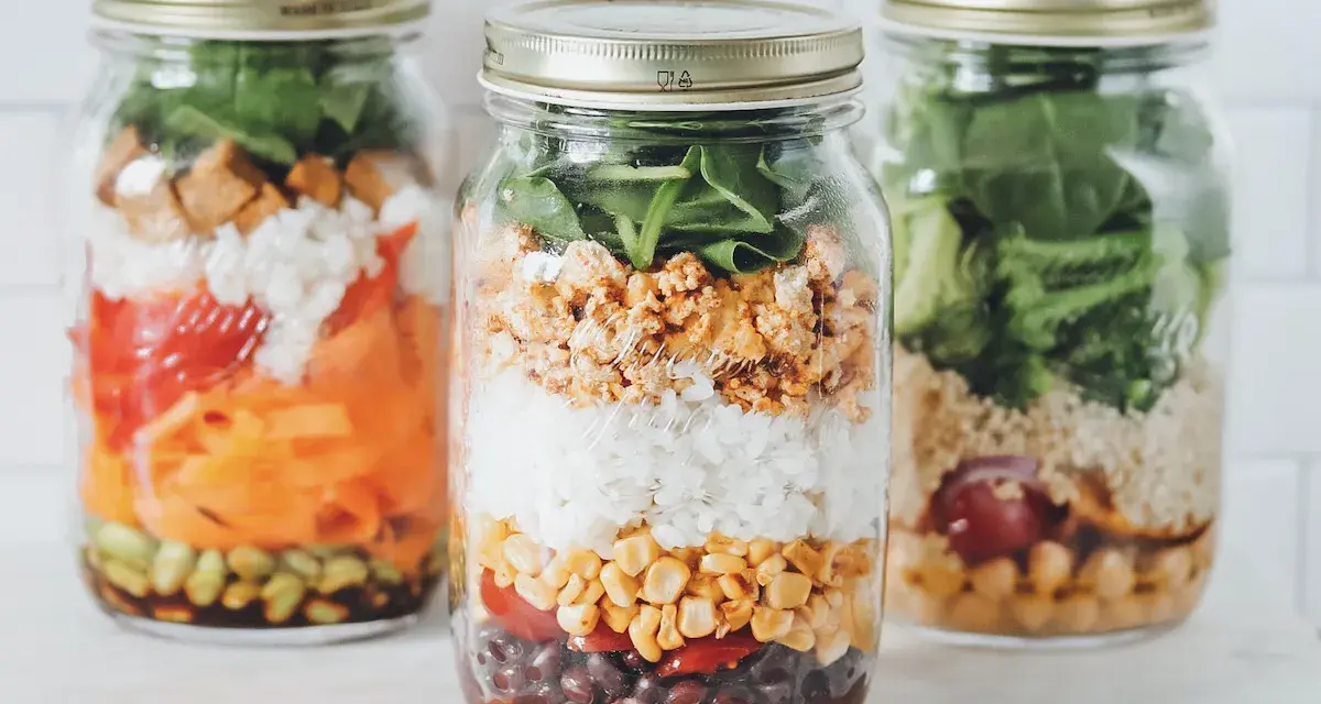 Drei bunte Schichtsalate im Glas, bereit für unterwegs. Jedes Glas ist ein kleines Kunstwerk mit Reis, Gemüse und Proteinen.