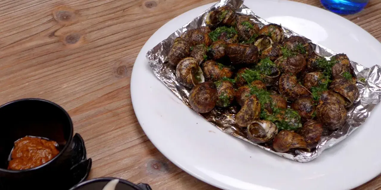 Caracoles con mantequilla y perejil, servidos en papel de aluminio sobre un plato blanco, junto a un cuenco con salsa.