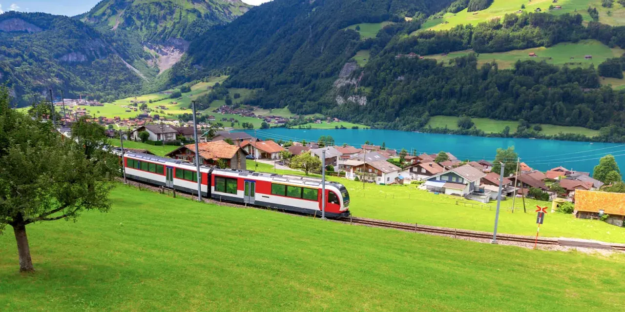 A red and white train travels along the **Golden Pass Line Lucerne to Interlaken**, passing a village by a turquoise lake, with lush green mountains in the background.