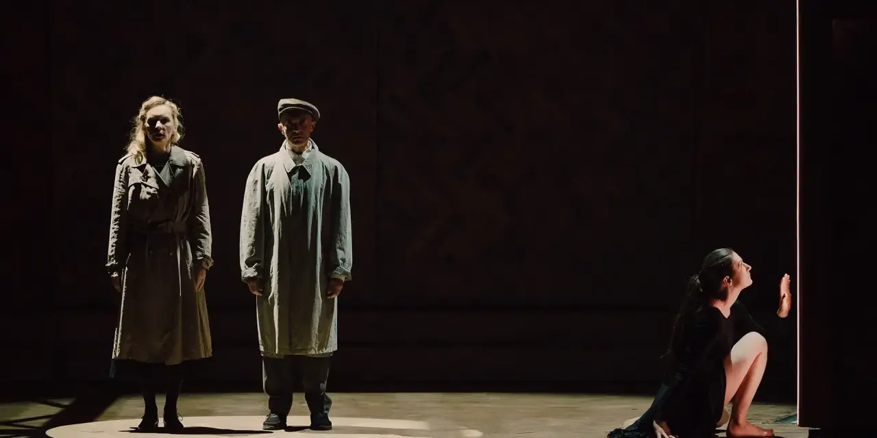 Magdalena Twardowska w scenie teatralnej. Dwie postacie w płaszczach stoją w świetle, obok nich kobieta w czarnej sukni klęczy przy pionowym świetle.