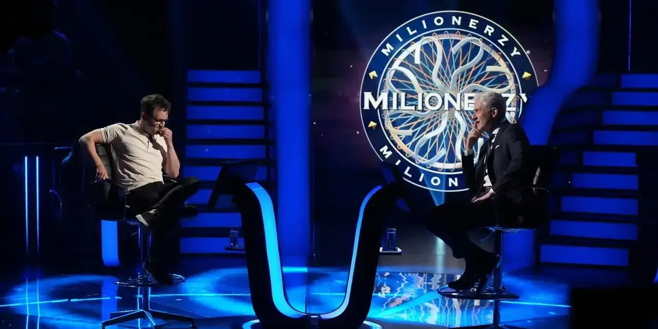 Dwóch mężczyzn siedzi naprzeciwko siebie na scenie "Milionerzy". Jeden w białej koszulce, drugi w garniturze.
