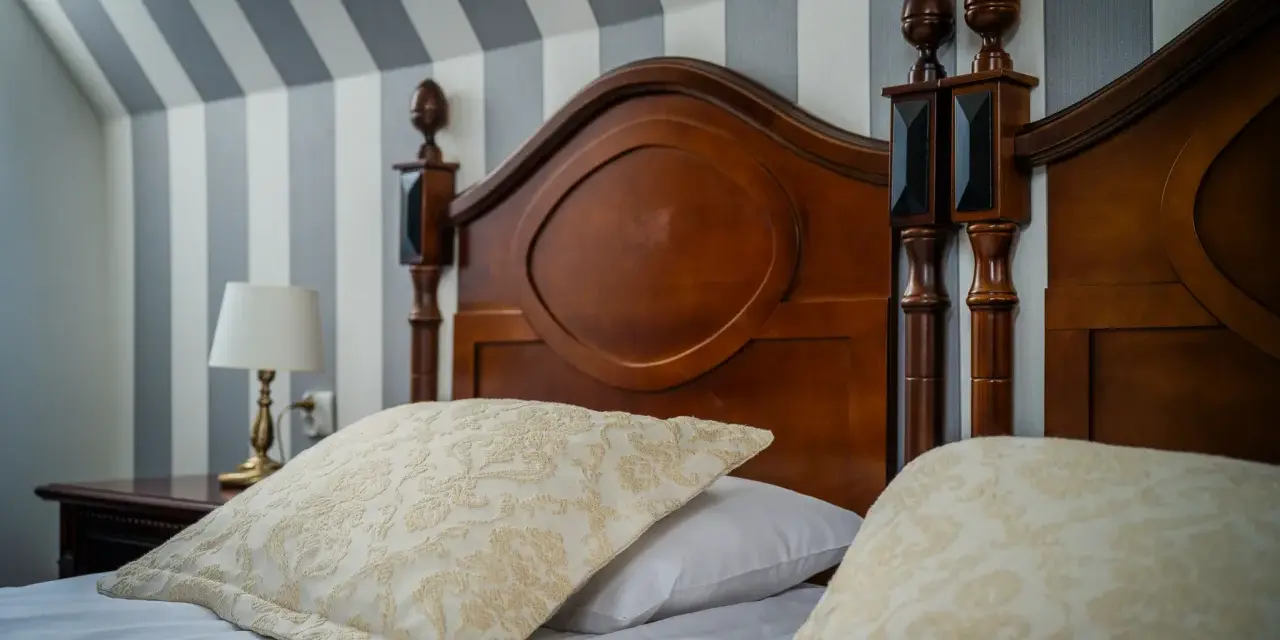 Eleganckie łóżko z drewnianym zagłówkiem i ozdobnymi poduszkami. Ciekawe, ile wynosi ryczałt za nocleg w takim miejscu?
