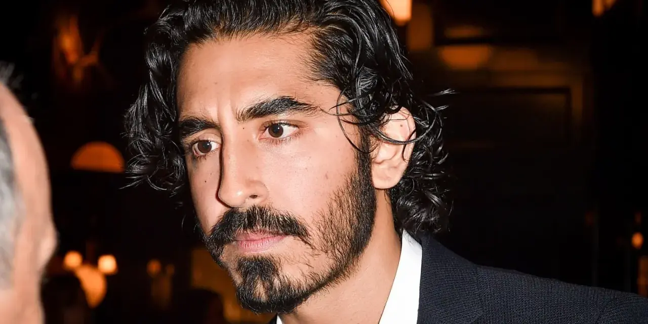 Primer plano de un hombre con cabello oscuro y rizado, y una barba poco poblada larga. Viste una camisa blanca y un traje oscuro.