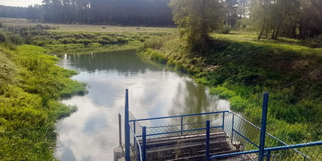 Spokojna rzeka otoczona zielenią, z widocznym pomostem i ogrodzeniem. Piękne zdjecia w wodzie odbijające niebo.