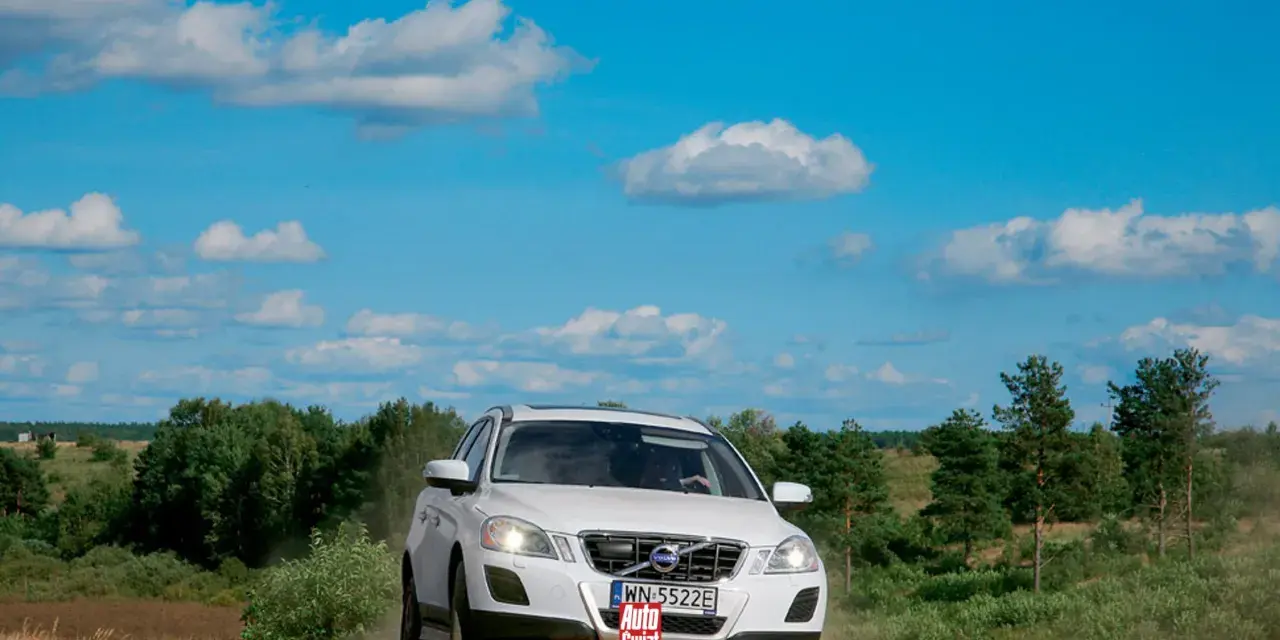 Białe Volvo XC60 T6 jedzie po piaszczystej drodze wśród zieleni, pod błękitnym niebem z chmurami.